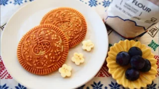 テフ粉使用のグルテンフリーパンケーキが2枚白い皿に並び、隣にはぶどうと花型のトッピング。背景にはカラフルなクロスとテフ粉の袋が置かれている