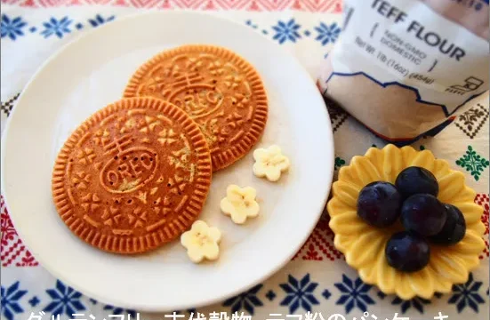 テフ粉使用のグルテンフリーパンケーキが2枚白い皿に並び、隣にはぶどうと花型のトッピング。背景にはカラフルなクロスとテフ粉の袋が置かれている