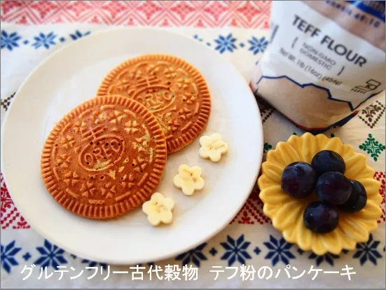テフ粉使用のグルテンフリーパンケーキが2枚白い皿に並び、隣にはぶどうと花型のトッピング。背景にはカラフルなクロスとテフ粉の袋が置かれている